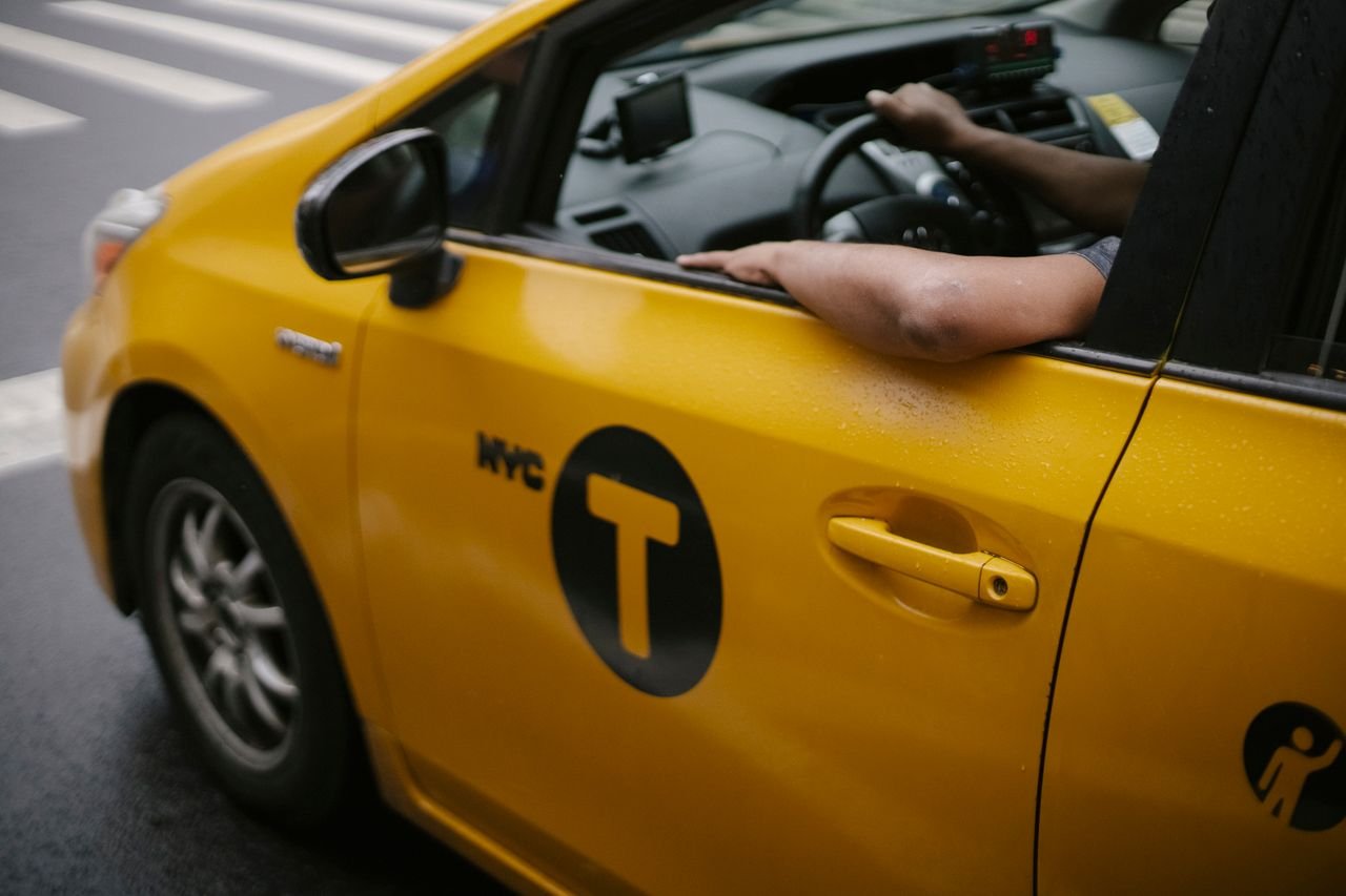 Unrecognizable man sitting in yellow taxi automobile and driving on roadway in town in daytime